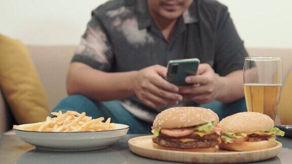 桌上汉堡包的4k视频在家沙发上用智能手机的年轻人不健康的生活理念