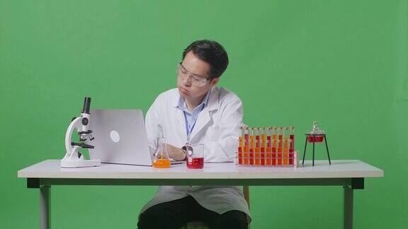 在绿屏背景的实验室里亚洲男科学家用试管做实验在笔记本电脑上打字然后在显微镜的桌子上头痛