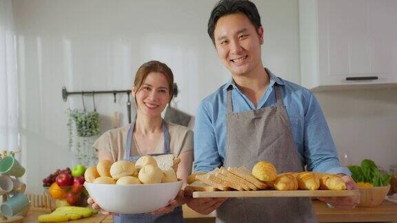 亚洲年轻夫妇的肖像拿着一碗面包看着相机漂亮的男男女女穿着围裙花时间在家里做早餐保健饮食和健康食品