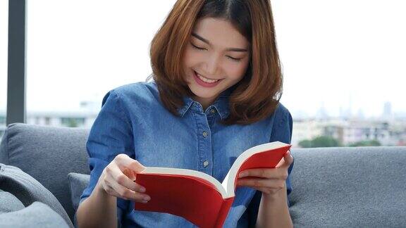 放松的亚洲女人坐在客厅的沙发上拿着书看书年轻女子放松读书打开书读书休闲心灵幸福美女人笑脸快乐的时候