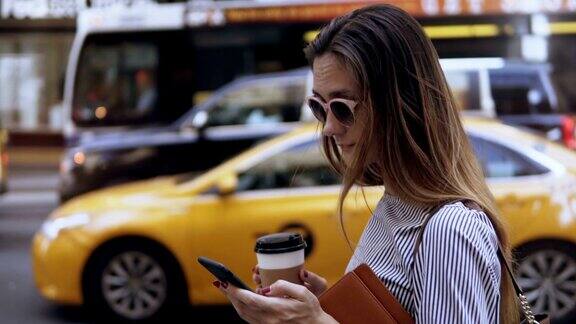 年轻漂亮的女商人站在路边喝着咖啡用着智能手机女性在等出租车