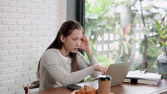 亚洲女人坐在咖啡馆的桌子上桌上放着笔记本电脑女人脸上带着严肃的表情假期坐着工作时手放在太阳穴上坐着感到头痛时