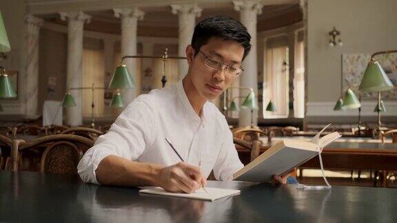 在镜头中一名年轻的亚洲男学生带着书在大学图书馆做学习笔记