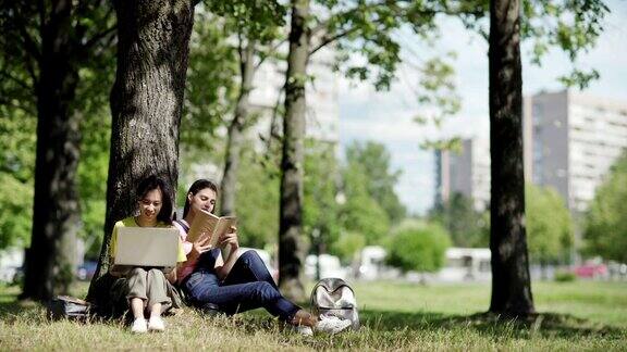 两个不同的女大学生坐在树下的公园在刮风的一天亚洲女孩在笔记本电脑上打字而她的白人朋友在看书