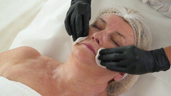俯视图美容师清洁高级妇女的皮肤与湿棉垫前面部护理程序抗衰老的治疗理念