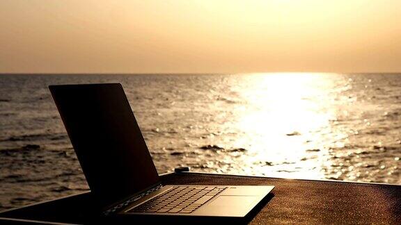 特写一个打开的笔记本电脑躺在阳光下的桌子上背景是日出或日落的海边