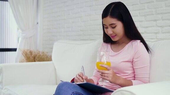 年轻快乐的亚洲女人写纸条喝橙汁坐在家里客厅的沙发上