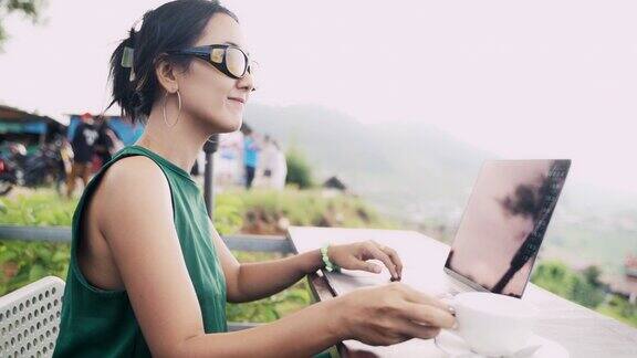 亚洲女性在山间自然中使用笔记本电脑工作喝着热咖啡