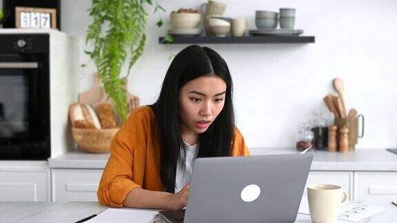 开朗感性的中国女孩穿着时髦的衣服自由职业者IT专家坐在厨房的桌子旁拿着笔记本电脑享受大利润庆祝胜利好消息用拳头打手势微笑