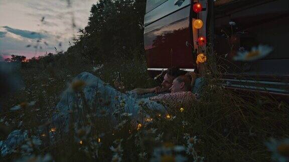 黄昏时分一对拿着智能手机的夫妇躺在草地上旁边是一辆停在草地上的露营车