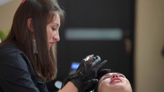 专业美容师纹身师在美容院为客户做永久眉妆的侧视图画像专注于年轻的高加索女人用微刀笔在皮肤上纹身