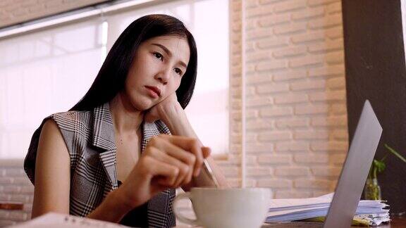 一个美丽的亚洲女人的特写肖像是用勺子搅拌咖啡来回显示无聊的迹象面包店老板坐在咖啡店用笔记本电脑做无聊的工作