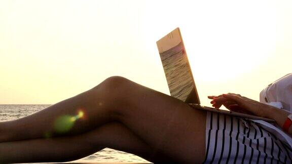 特写穿着短裤的女性双腿年轻女子躺在躺椅上在海边的沙滩上在日落或日出时使用笔记本电脑自由职业者远程工作暑假在海上