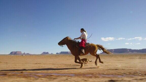 在一个晴朗晴朗的日子里一个十几岁的印第安女孩穿着传统的纳瓦霍服装骑着她的马疾驰在纪念碑谷沙漠和她的宠物狗