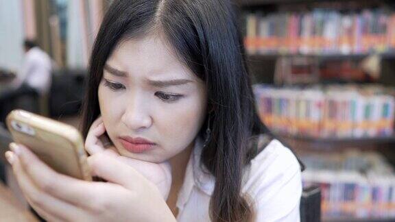 亚洲女学生使用智能手机聊天坐在图书馆靠近书架的书桌旁