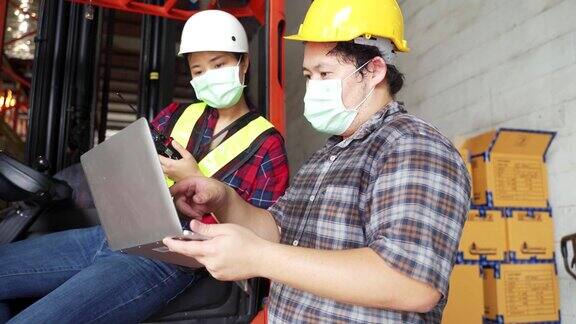 亚洲女工人坐在仓库的铲车里新冠肺炎疫情期间男女工程师戴安全帽、背心和面罩检查工厂的储物箱包裹