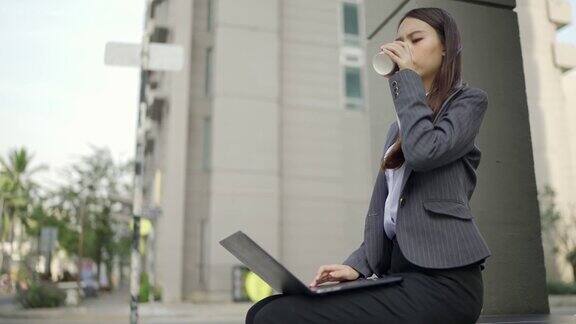 视频缓慢一个漂亮的白皮肤亚洲女人穿着黑色西装坐在办公大楼前在城市的路边使用笔记本电脑