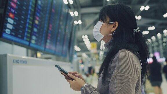 一名亚洲女性戴着医用口罩用智能手机和手表查看机场的航班时刻表