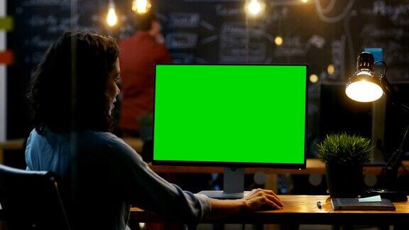 美丽的女办公室职员在她的桌面工作在一个模拟的绿屏个人电脑过肩镜头她的同事在后台工作晚上创意办公室