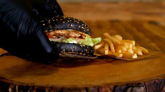 戴上手套拿起一个多汁的黑色汉堡里面有肉片和蔬菜不健康的快餐薯条还留在木板上