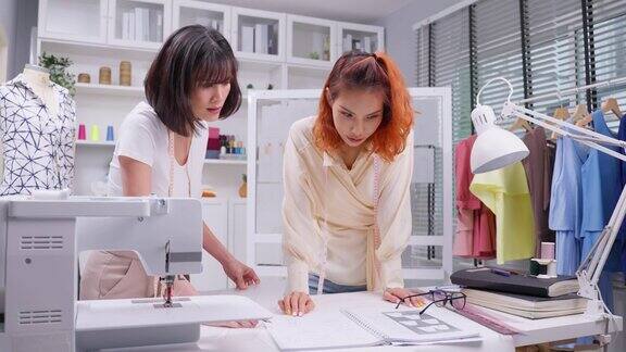 亚洲两个裁缝妇女工作设计服装在裁缝工作室迷人的年轻女时装设计师女裁缝在工作室使用笔记本电脑创作新的图案集合