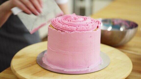 女性糕点厨师使用糕点刮板和旋转蛋糕架光滑的粉红色奶油霜手工蛋糕的特写跟踪右镜头