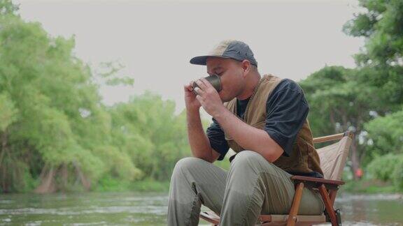 快乐的亚洲人早上坐在河边喝咖啡在大自然中露营他感到放松和清爽休闲旅行和户外活动的生活方式