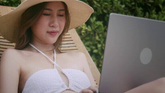 一位亚洲女性躺在泳池边的椅子上用笔记本电脑工作