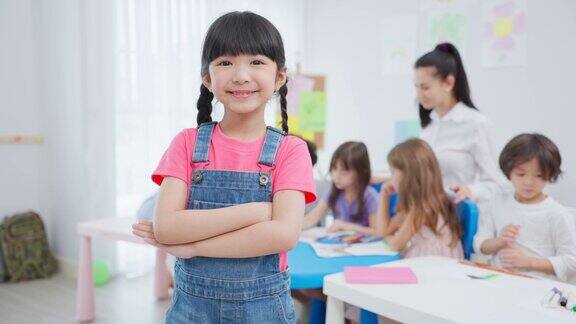 亚洲小女生的肖像幸福地站在教室里年轻可爱的学龄前儿童交叉手臂看着镜头而女教师在幼儿园教学活动