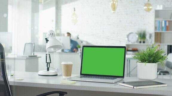 绿色屏幕的笔记本电脑站在桌子上没有人在上面工作背景里一个男人走进来坐在他的工作场所开始工作