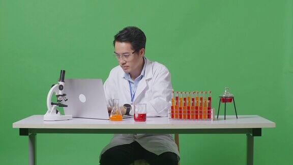 在绿屏背景实验室里亚裔男科学家用试管做实验尖叫着庆祝进球在显微镜下的桌子上用笔记本电脑打字