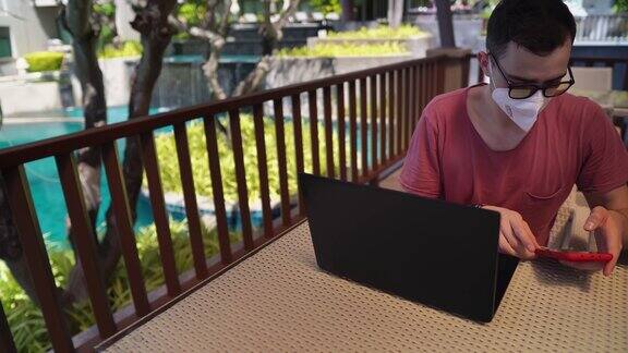 一名男子坐在酒店大堂使用智能手机面前放着一台打开的笔记本电脑