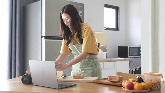 年轻的亚洲妇女通过笔记本电脑学习在线烹饪课程制作三明治人们健康的生活方式