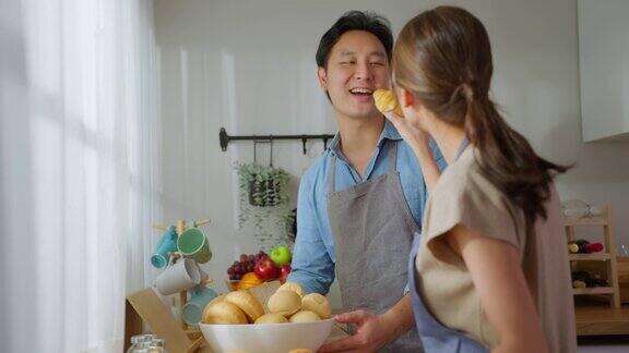 亚洲年轻女性拿着面包在厨房给男性有魅力的男女夫妇系着围裙花时间在家里做饭吃早餐饮食与健康食品理念