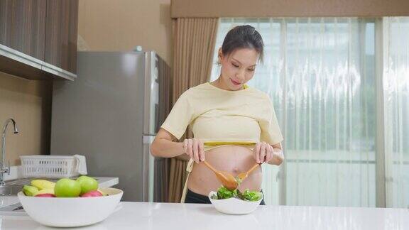 亚洲美丽的孕妇在家里厨房烹饪健康的食物迷人的怀孕女孩站在厨房的室内感到快乐享受在碗绿色蔬菜沙拉的健康在家里