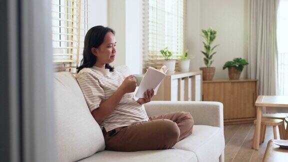 年轻的亚洲妇女喜欢在早上坐在客厅家里的沙发上看书和喝咖啡周末读一些喜欢的书放松一下