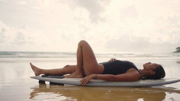 4K迷人的亚洲女子泳装躺在冲浪板在热带海滩在夏天的日落