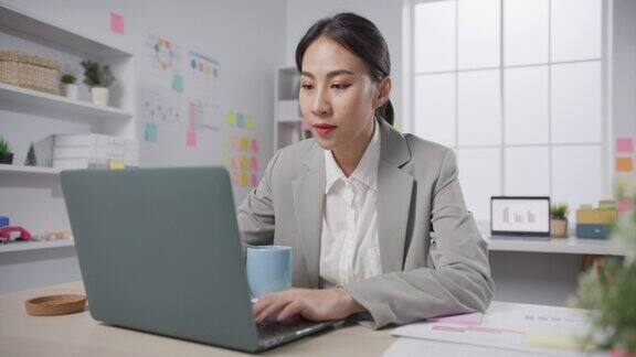 年轻的亚洲开朗的专业女商人财务顾问顾问坐在桌子上笔记本电脑喝咖啡在现代办公室在线工作