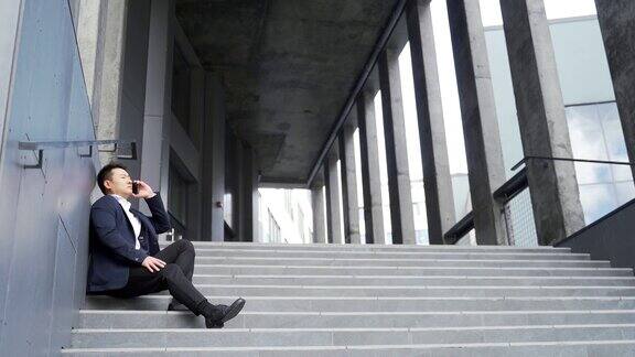 年轻的亚洲人沮丧地坐在楼梯上的街道附近的办公商业中心大楼