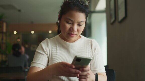 微笑开朗的亚洲女性放松手使用智能手机上网冲浪社交媒体而坐在咖啡馆白天