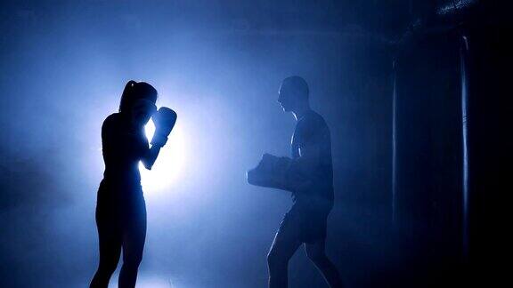 在烟雾缭绕的体育馆一名女拳击手戴着拳击手套击打拳击袋的剪影
