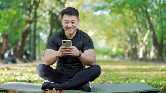 快乐的亚洲人穿着运动服坐在垫子上在城市公园浏览智能手机图为一名英俊的男子正在微笑着阅读在线聊天参与者的留言