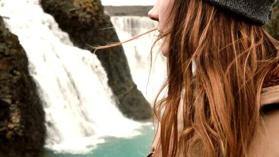 近距离观看年轻女子站在冰岛一个强大的瀑布附近头发随风摆动