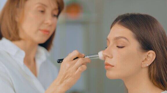 美丽的年轻女子在美容工作室化妆化妆师用刷子把粉涂在脸上肖像的观点
