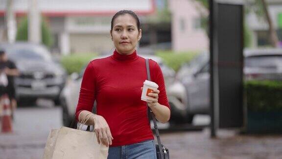 一名亚洲妇女背着购物袋喝着咖啡沿着马路快乐地四处张望
