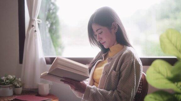 快乐的年轻亚洲女子在家中明亮的客厅室内靠窗看书有时间放松