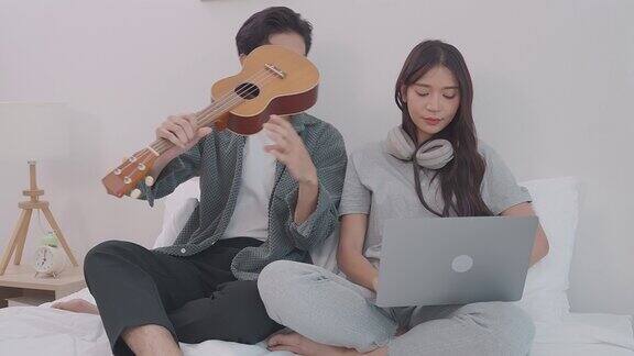 年轻迷人的亚洲夫妇坐在一起看笔记本电脑在卧室里男人和女人在家里的床上用笔记本电脑在线弹吉他爱情和夫妻关系