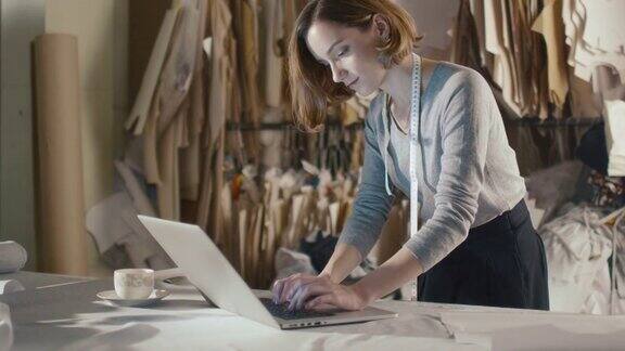 一位女服装设计师倚靠在工作室的桌子上正在用笔记本电脑工作