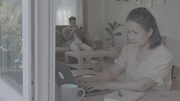 一名亚洲女学生在家里的客厅里上网学习时一边在笔记本电脑键盘上打字她的朋友就坐在旁边的沙发上