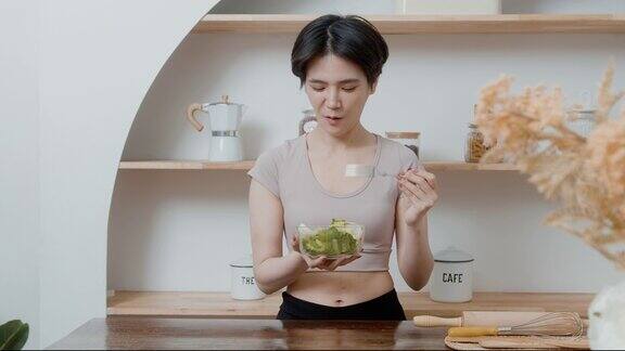 一位穿着运动服的亚洲年轻女子在家里的厨房里吃蔬菜沙拉